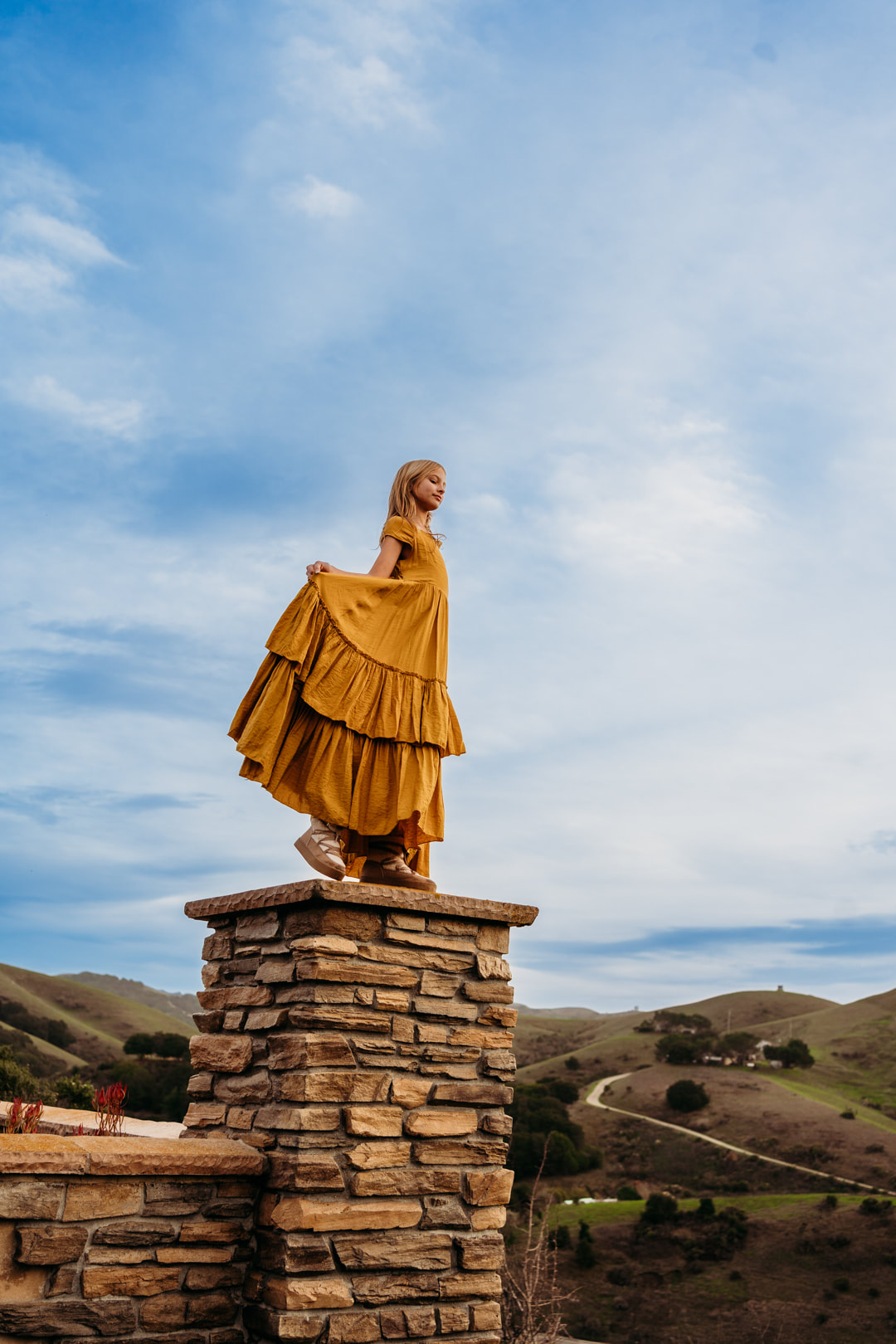 An 11 year old girl stands on a wall, holding the skirt of her ruffly dress against a blue sky.