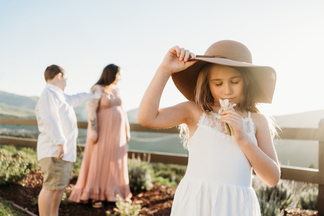 An 8 year old girl in a white dress holds her hat while smelling a flower.  In the background her mother and brother look out over the Danville hills.
