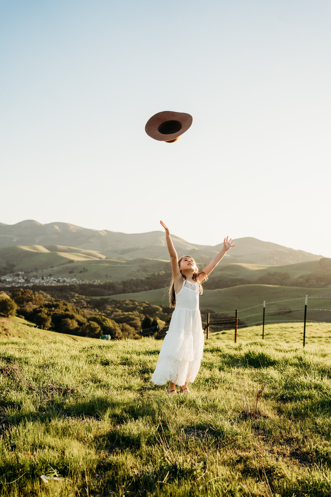A girl in a white dress throws her hat into the air on a grassy patch of the Danville hills.