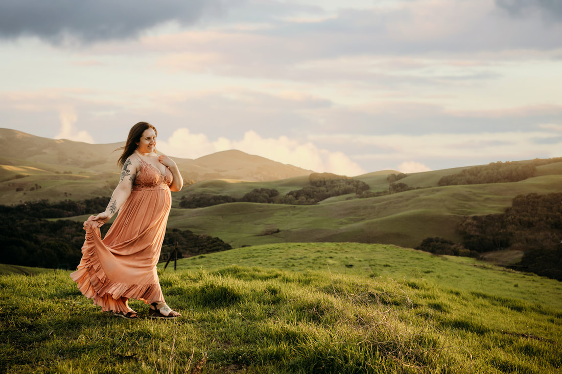 A woman in a pink dress gazes off towards the sunset while standing in the green hills above Danville.
