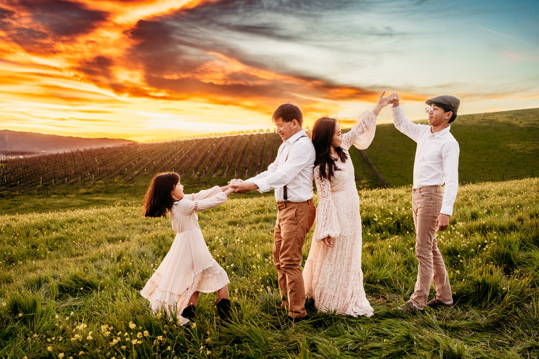 A family with two children dance on a hillside of flowers at sunset with a Pleasanton vineyard in the background.