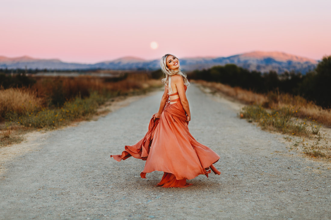 A high school senior girl twirls in a pink dress in front of the Livermore hills