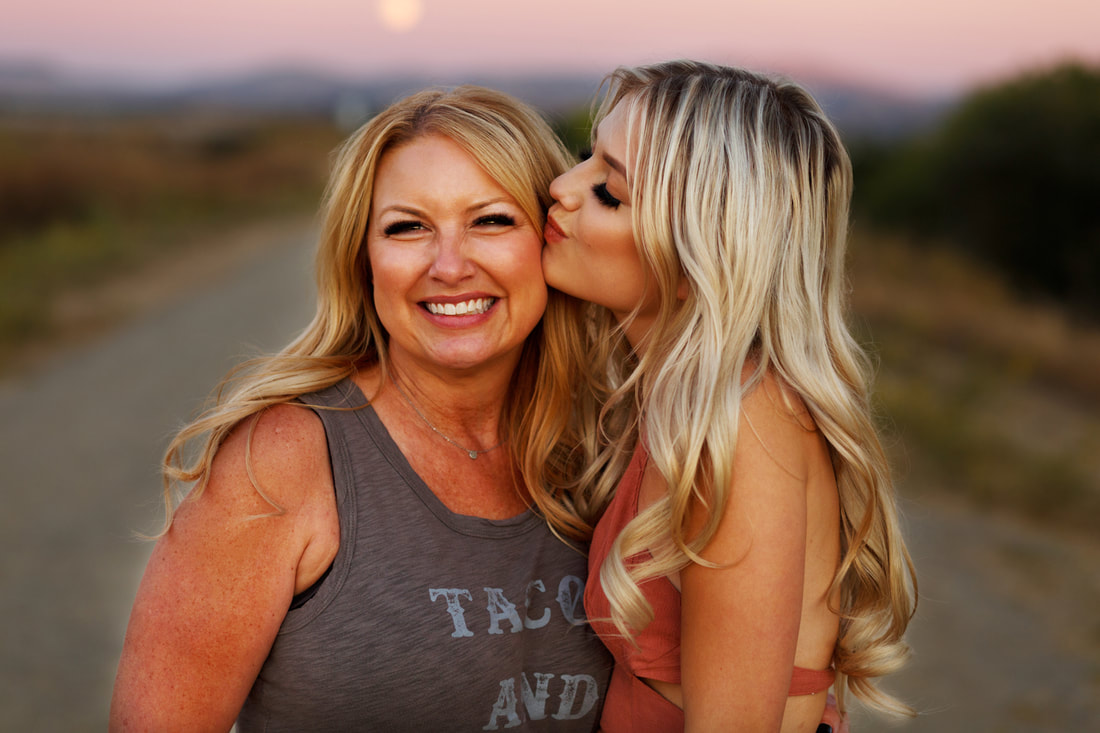 A high school senior girl kisses her mother's cheek with the pink sunset skies behind them