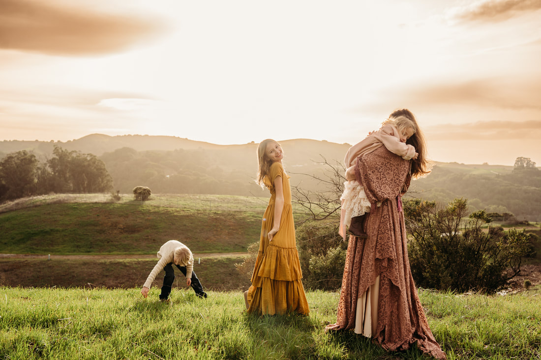A family portrait of a boy reaching down to the grass, a girl looking back at the camera, and a mother holding a toddler with the sun setting over the Pleasanton hills.