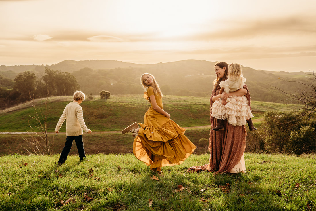 A candid family portrait with a boy looking away, a girl twirling in a yellow dress, while a mother watches and holds a toddler in the Pleasanton hills.