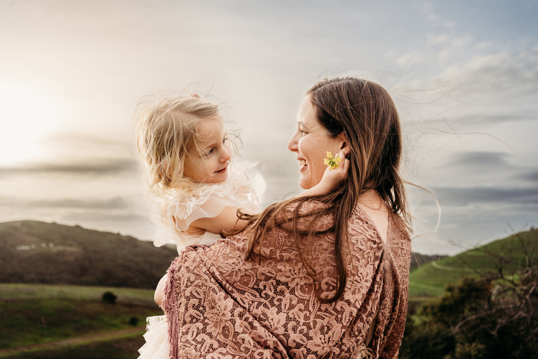 A mother holds her toddler daughter with the sun setting over the hills in Pleasanton while the girl places a flower in her hair.