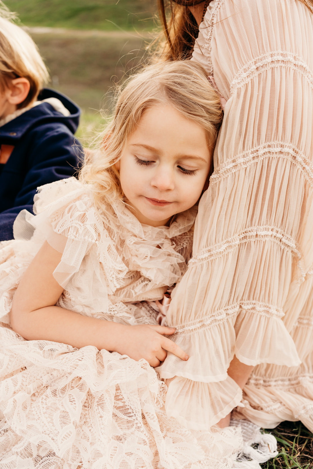 A four year old girl in a pink ruffly dress sleeps while holding her mother, sitting in the Pleasanton hills.