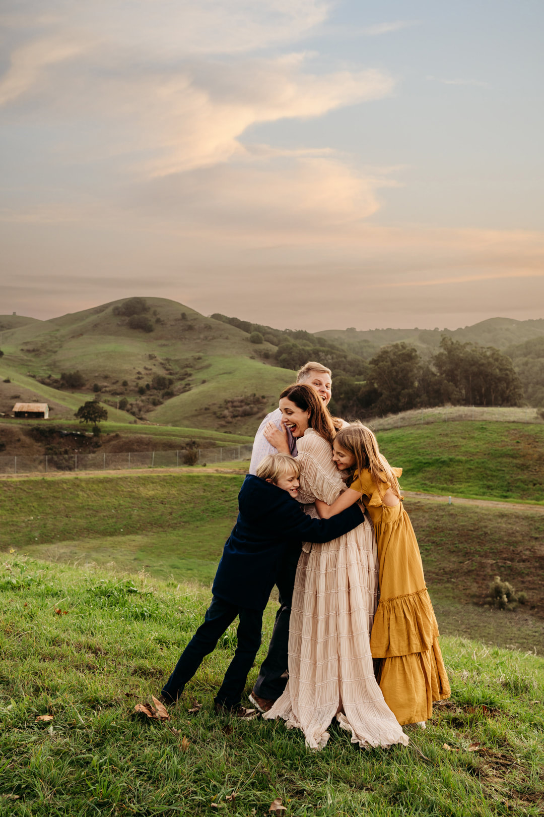 A family hugs and laughs with the rolling hills of Pleasanton and a small barn in the background.