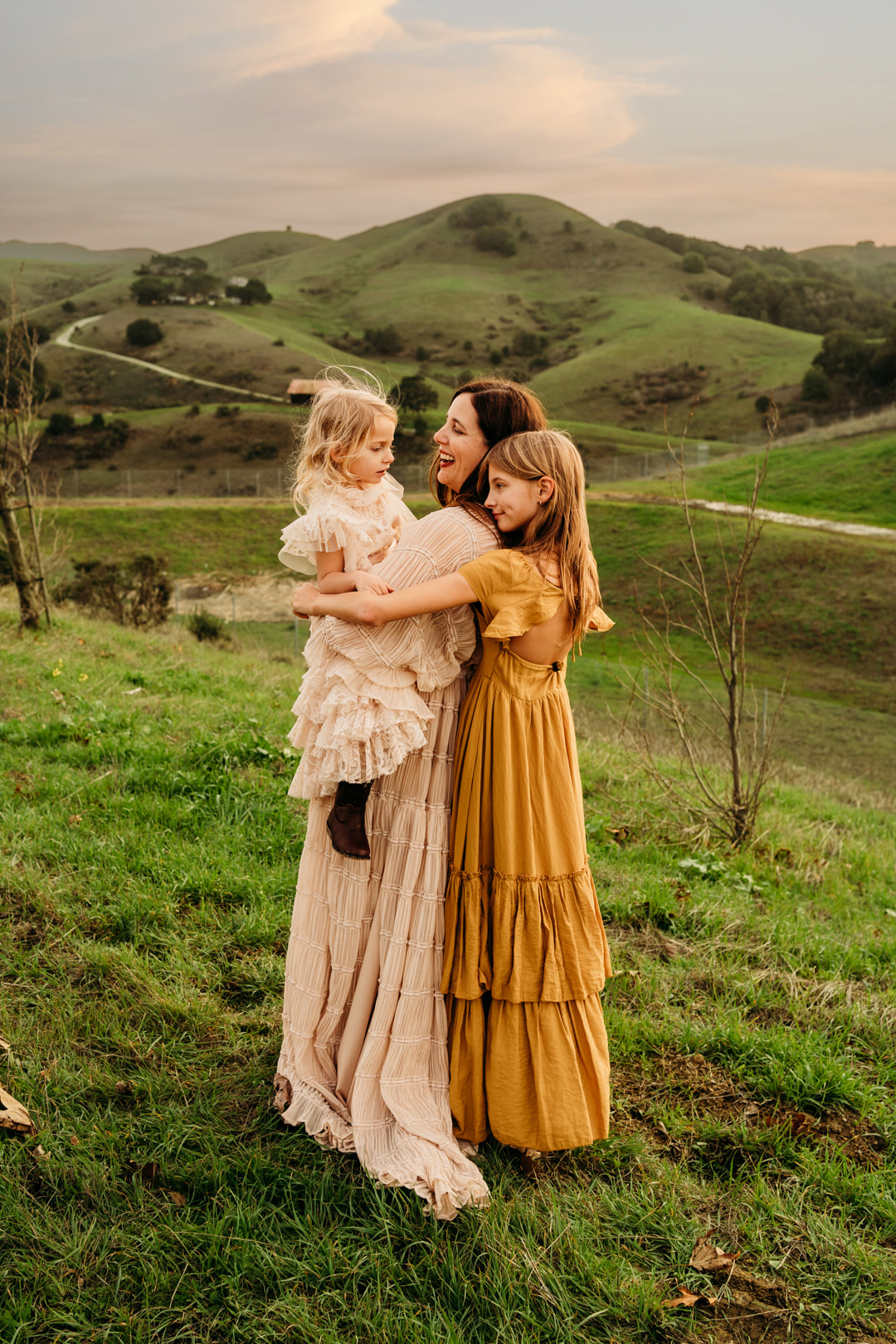 A group hug featuring a mother and her two daughters with a barn and the rolling hills of Pleasanton in the background.